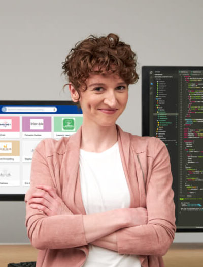 Coder standing in front of computer screens and smiling with her arms crossed. The screens behind her are displaying code and the Partner Portal interface.